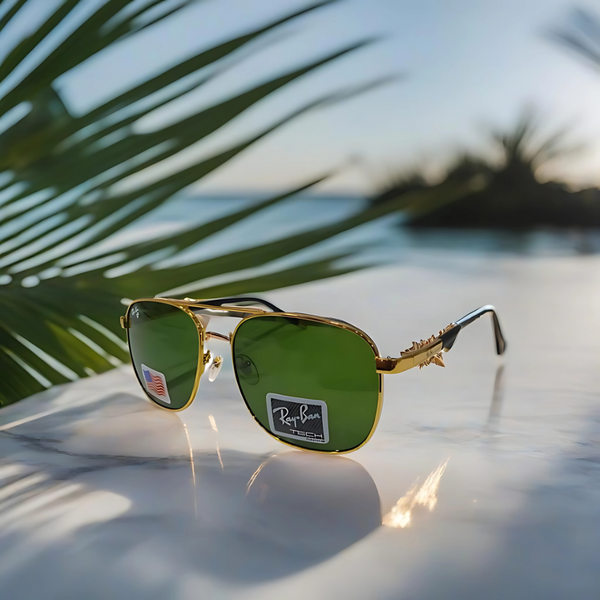 A pair of Rayban Pilot Aviator sunglasses with green lenses and a gold frame, displayed on a surface with a palm tree in the background.