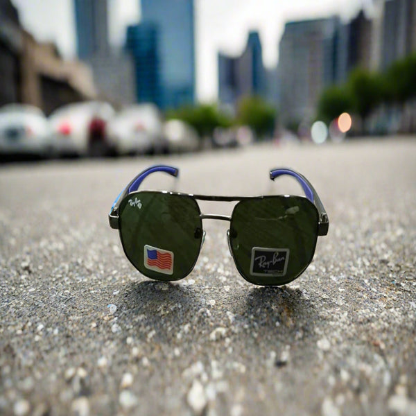 A pair of Ray-Ban sunglasses with green lenses and a metallic frame, placed on a reflective surface with a cityscape in the background.