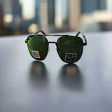 A pair of Rayban Pilot Aviator sunglasses with green lenses and a black frame, displayed on a reflective surface with a blurred cityscape background.