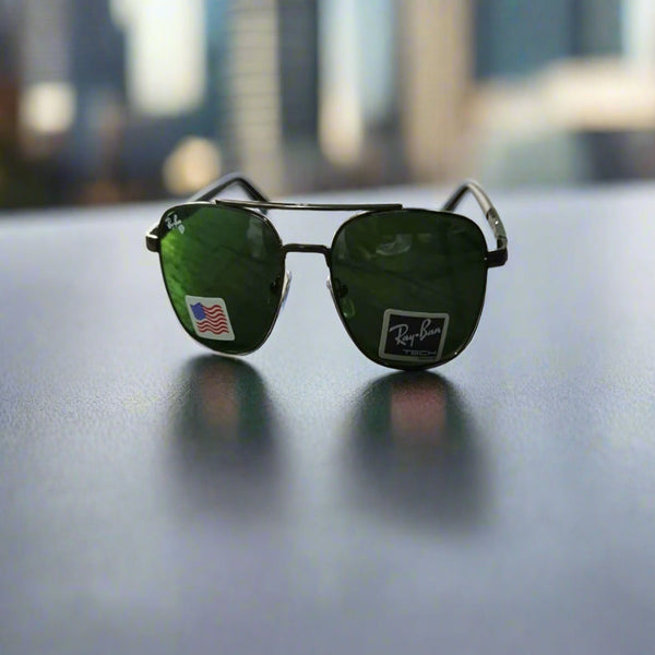 A pair of Rayban Pilot Aviator sunglasses with green lenses and a black frame, displayed on a reflective surface with a blurred cityscape background.