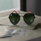 Aviator sunglasses with green lenses and red frames on a textured surface.